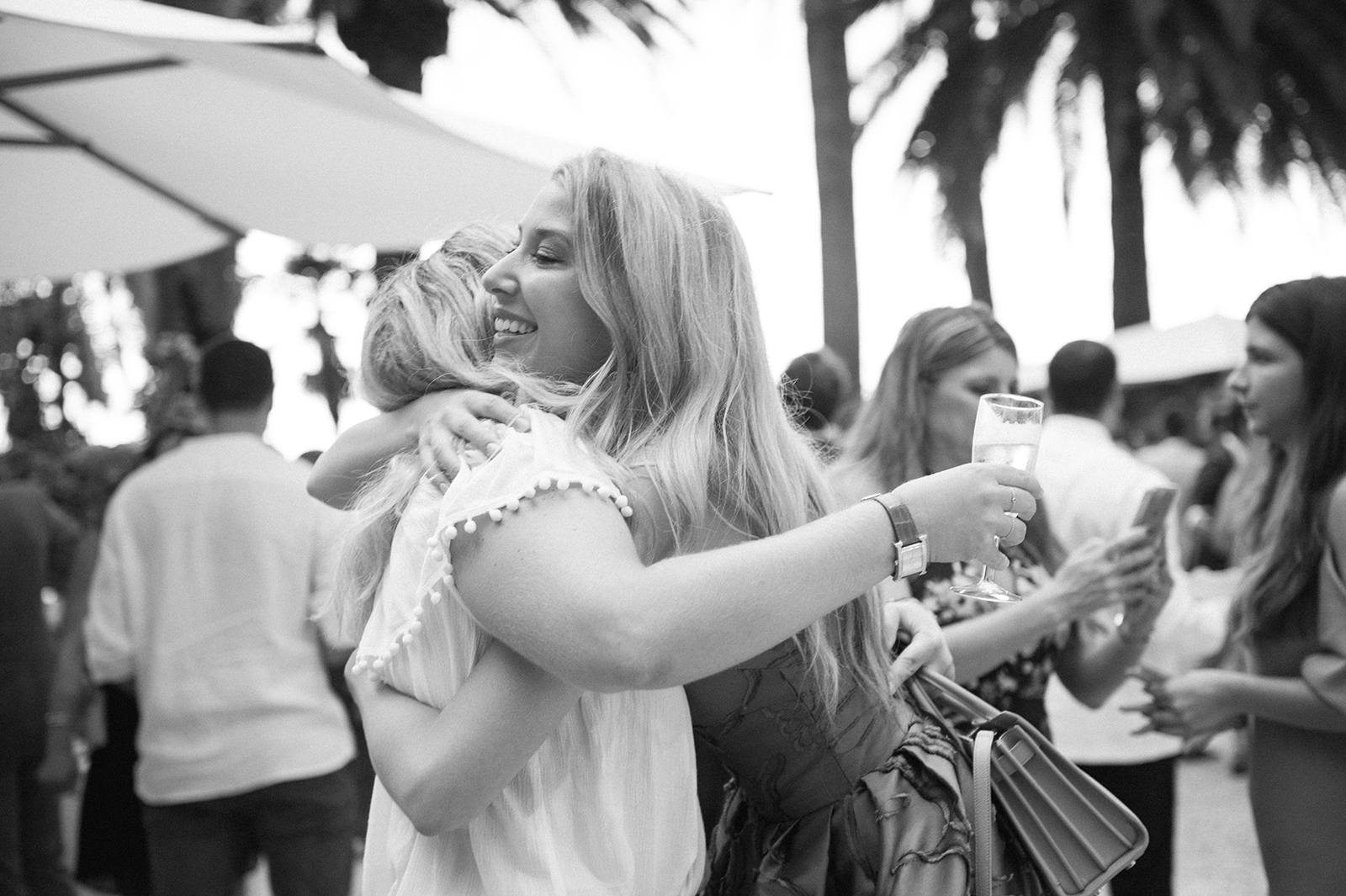 Black and white image of two women hugging and smiling while holding champagne at an outdoor wedding