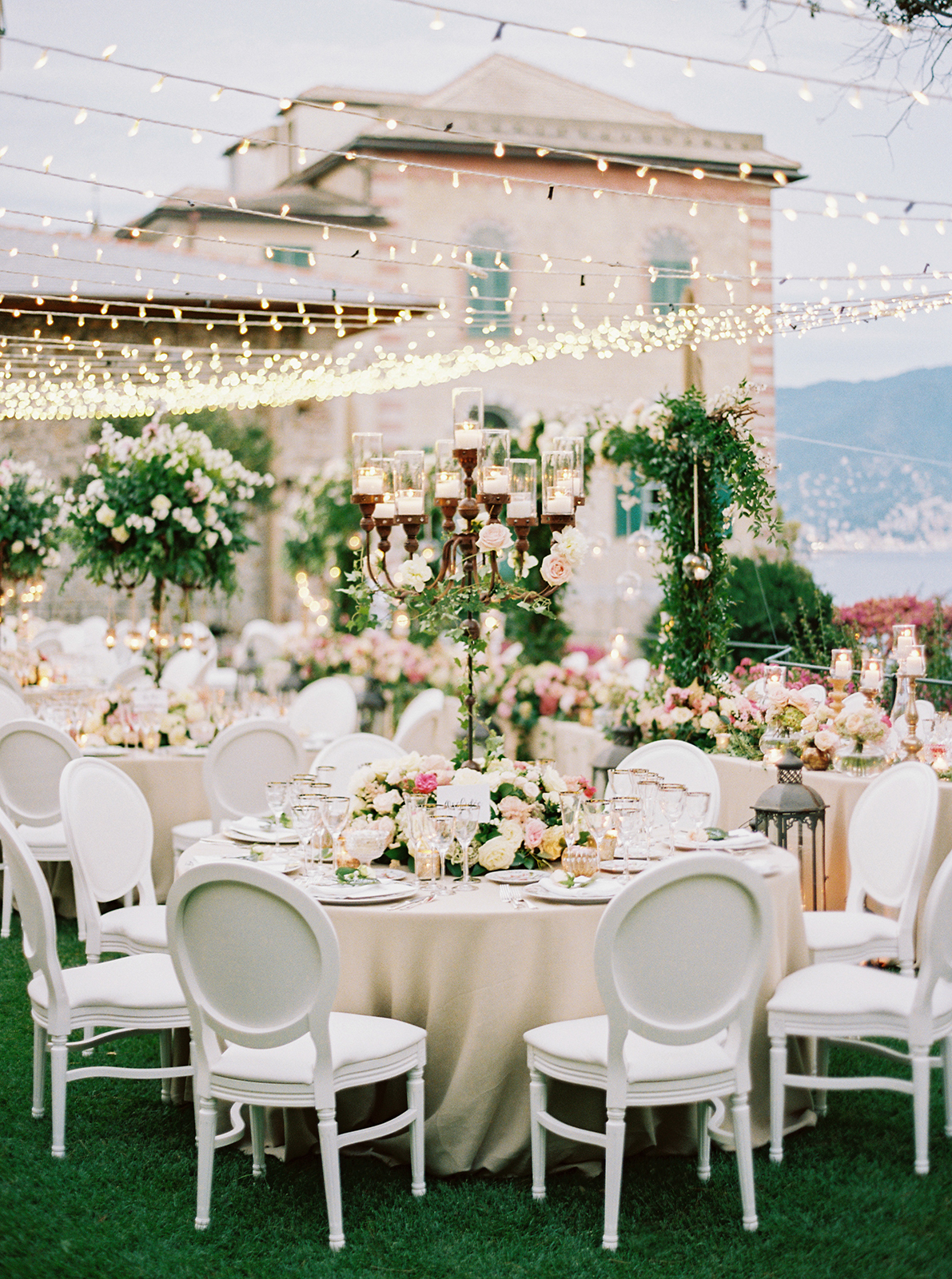 Elegant outdoor wedding reception with round tables, white chairs, floral centerpieces, and string lights at dusk