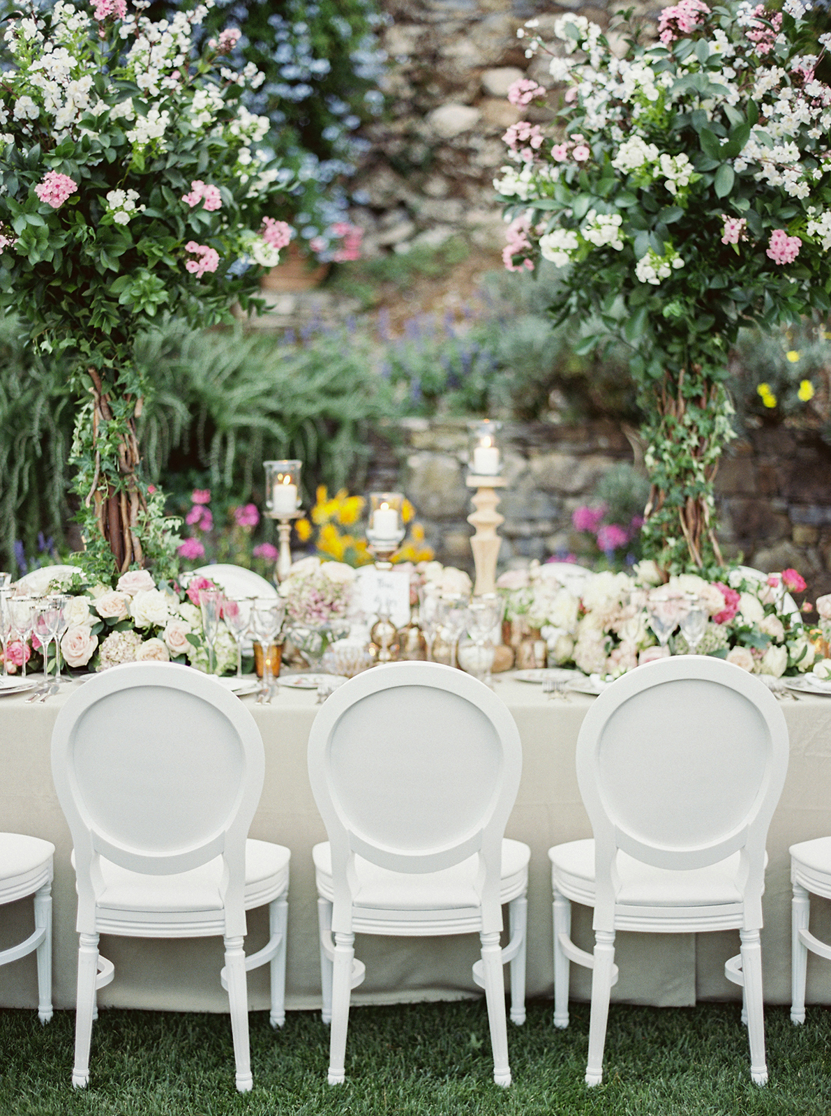Round wedding reception tables with tall floral centerpieces and overhead string lights in an outdoor garden setting