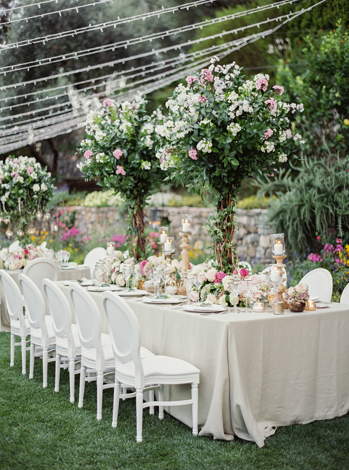 Garden wedding head table styled with white chairs, abundant floral arrangements, and candlelit details