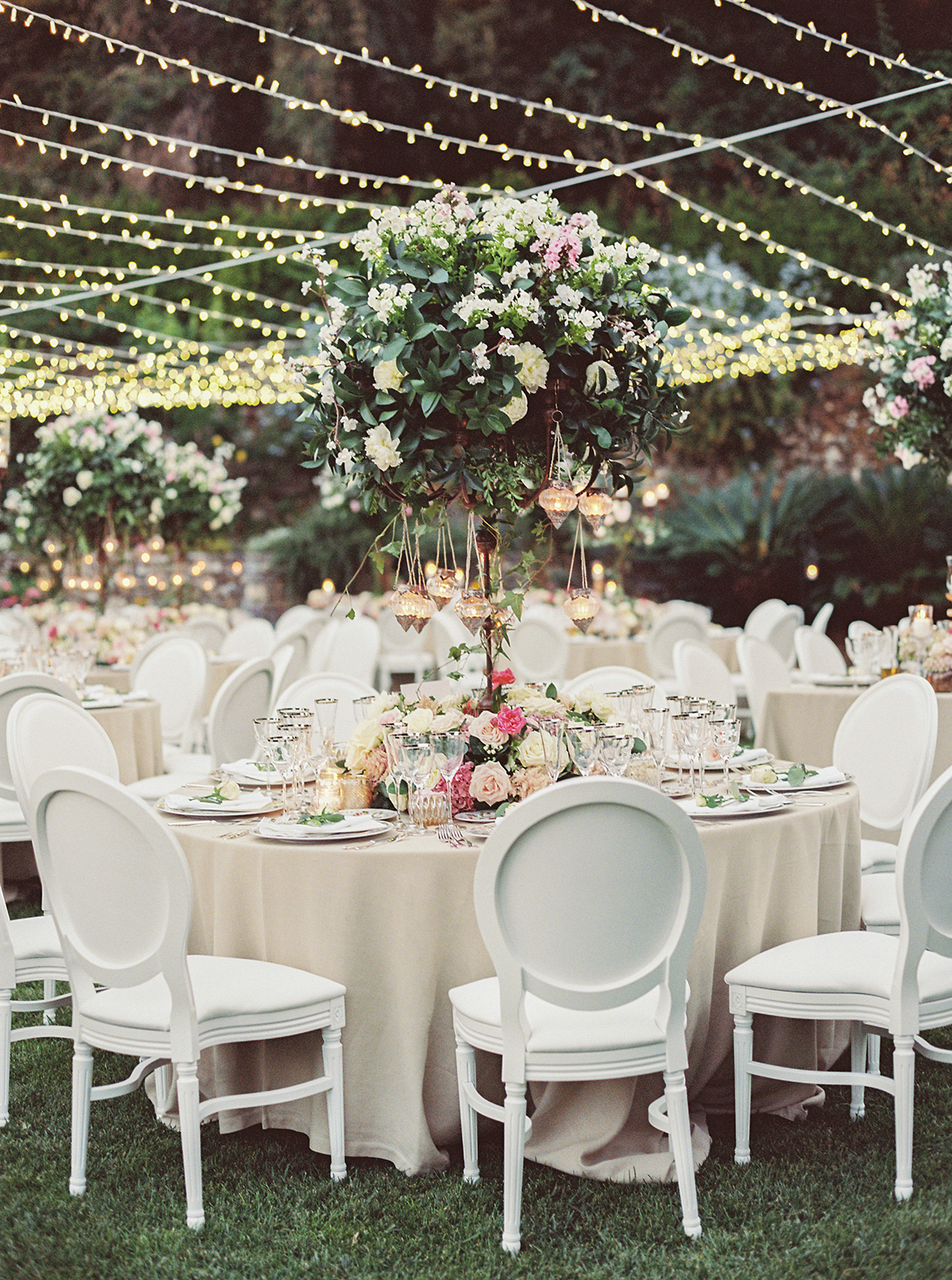 Outdoor wedding reception with round tables, hanging lights, and floral centerpieces under the trees