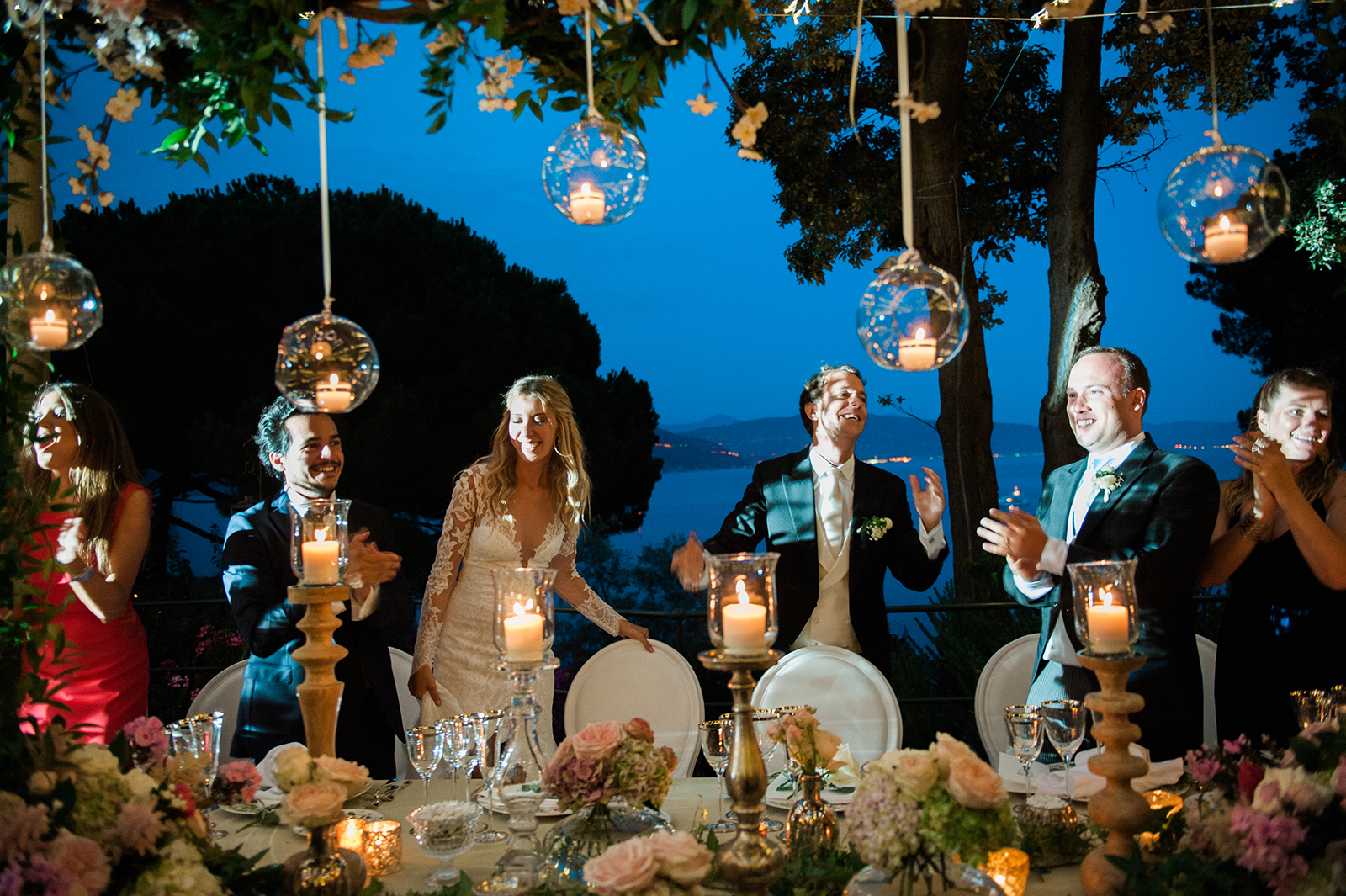 Bride and groom standing and applauding during an outdoor wedding dinner under hanging candle lanterns at night in la Cervara venue in Portofino