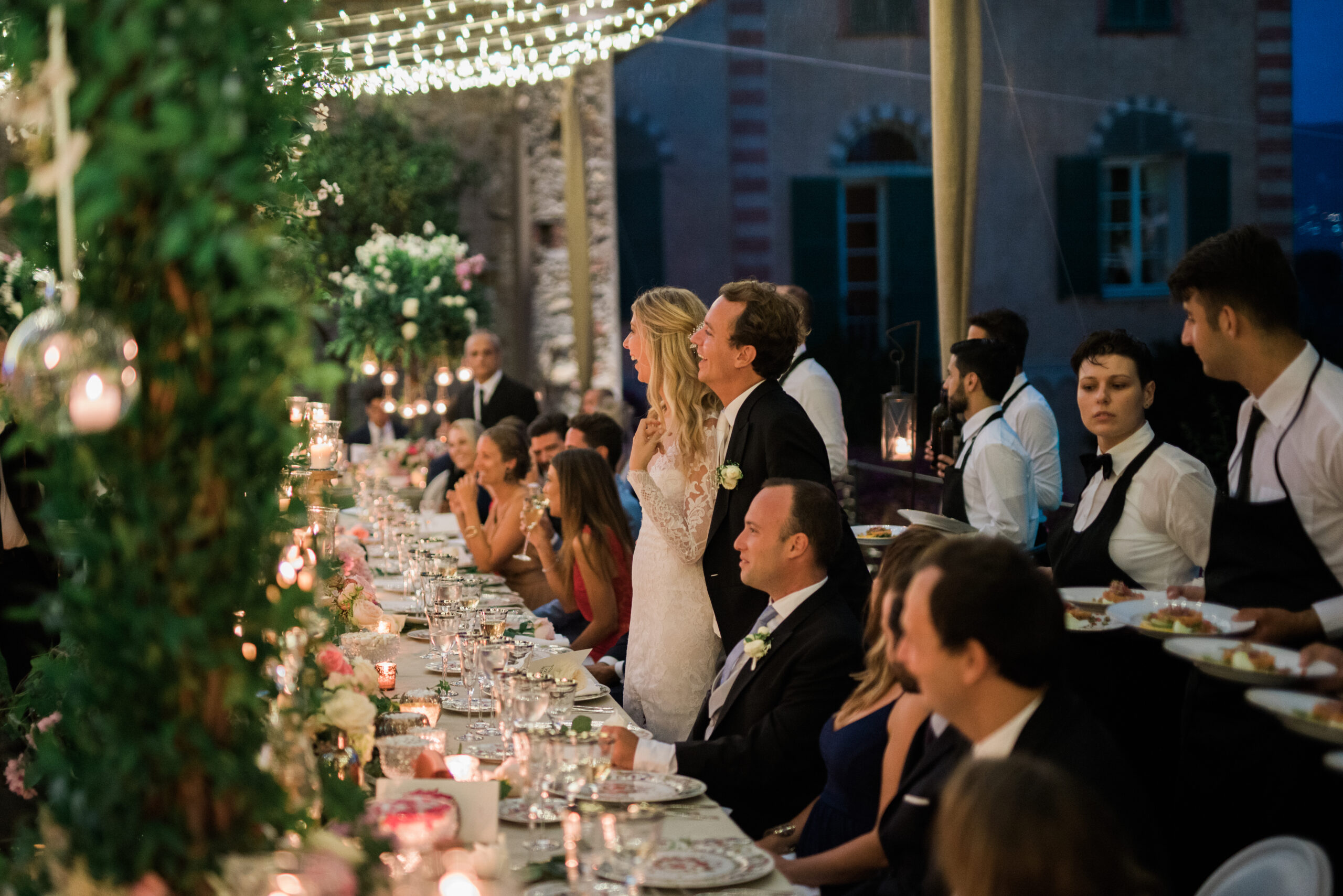 Long outdoor wedding dinner with guests seated at a candlelit table beneath string lights and greenery