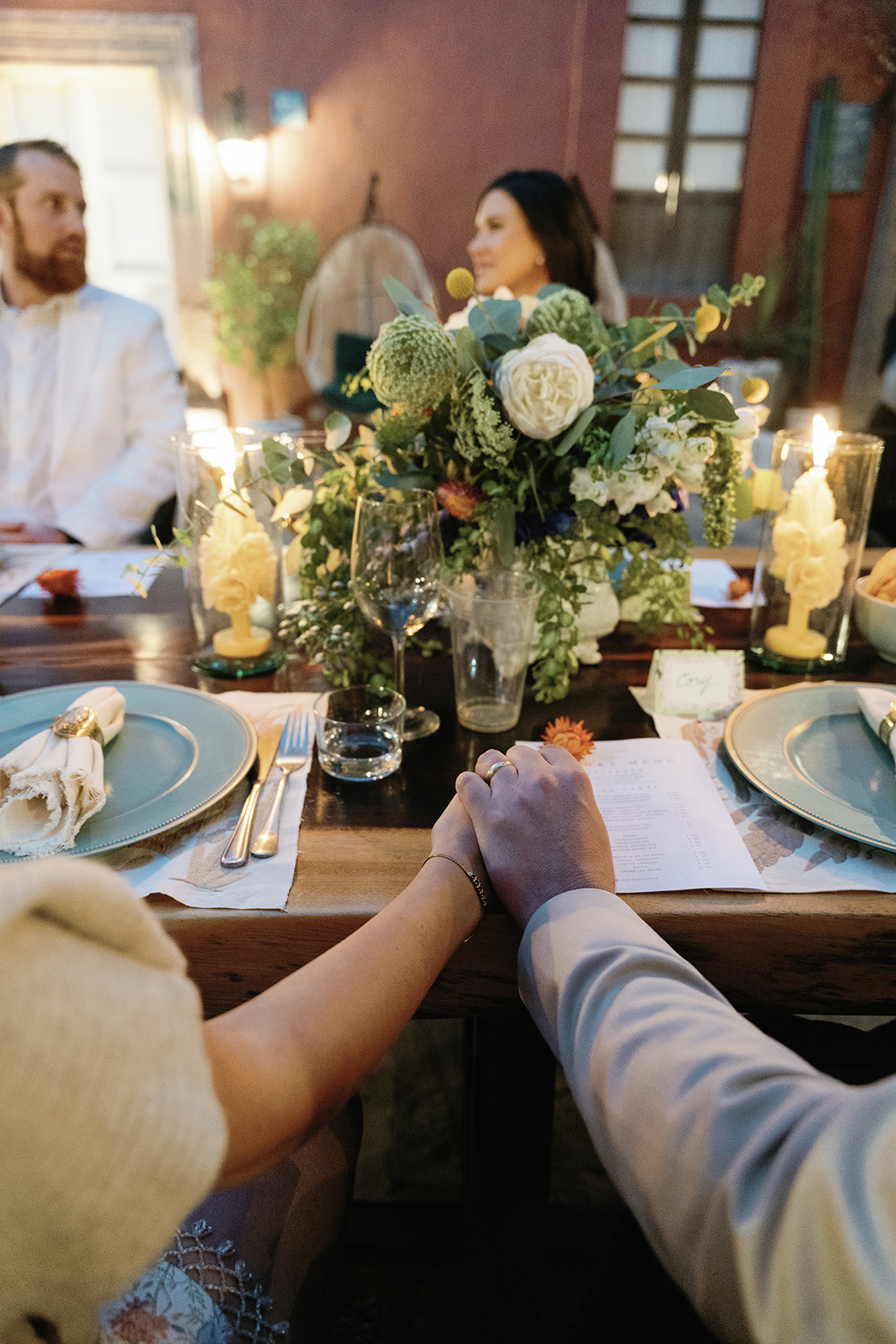 Wedding couple holding hands across dinner table surrounded by flowers and candles at Casa Hoyos