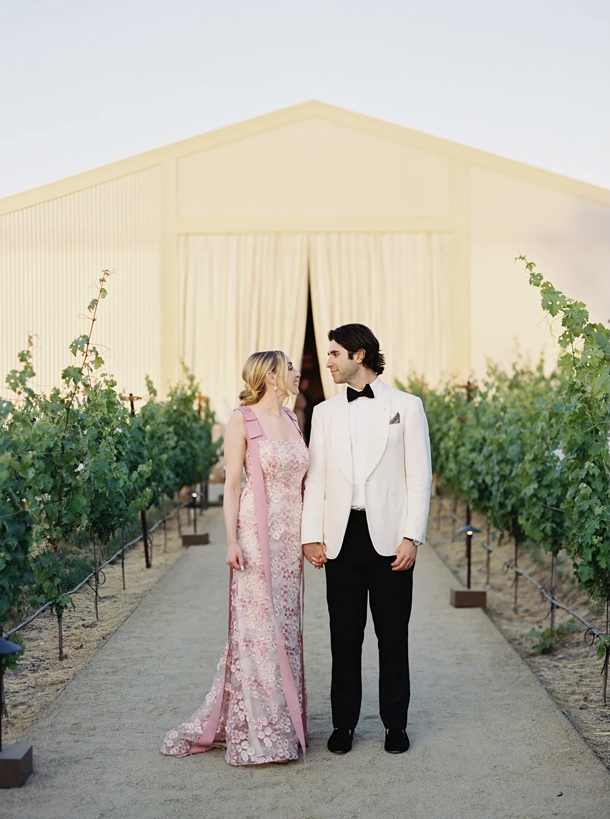 Bride in pink floral gown and groom in white tuxedo walking through vineyard path at Montage Healdsburg