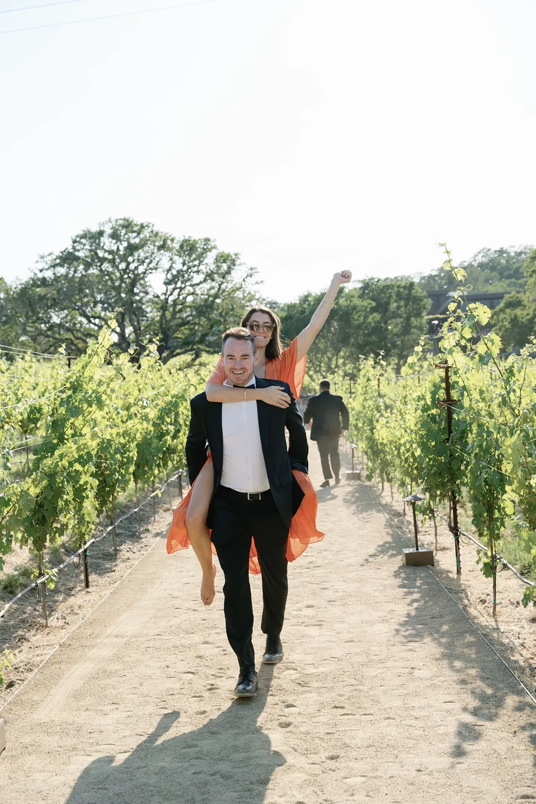 Wedding guests walking through vineyard rows, with one guest carrying another playfully along a sunlit path