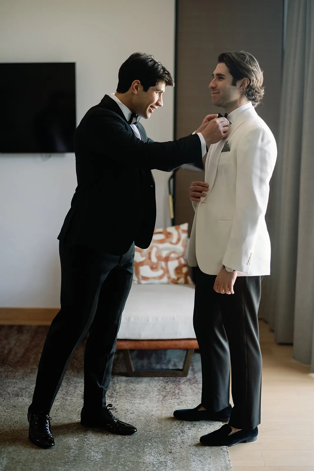 Best man adjusting the groom’s bow tie while they get ready together in the suite