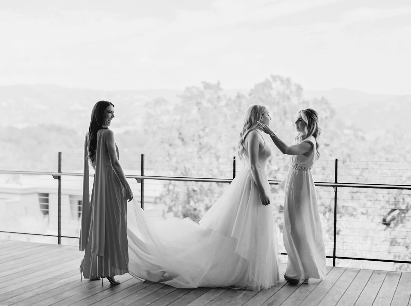Black-and-white image of a bride standing on a wooden terrace at Montage Healdsburg as two women adjust her hair and gown, with vineyard hills in the background