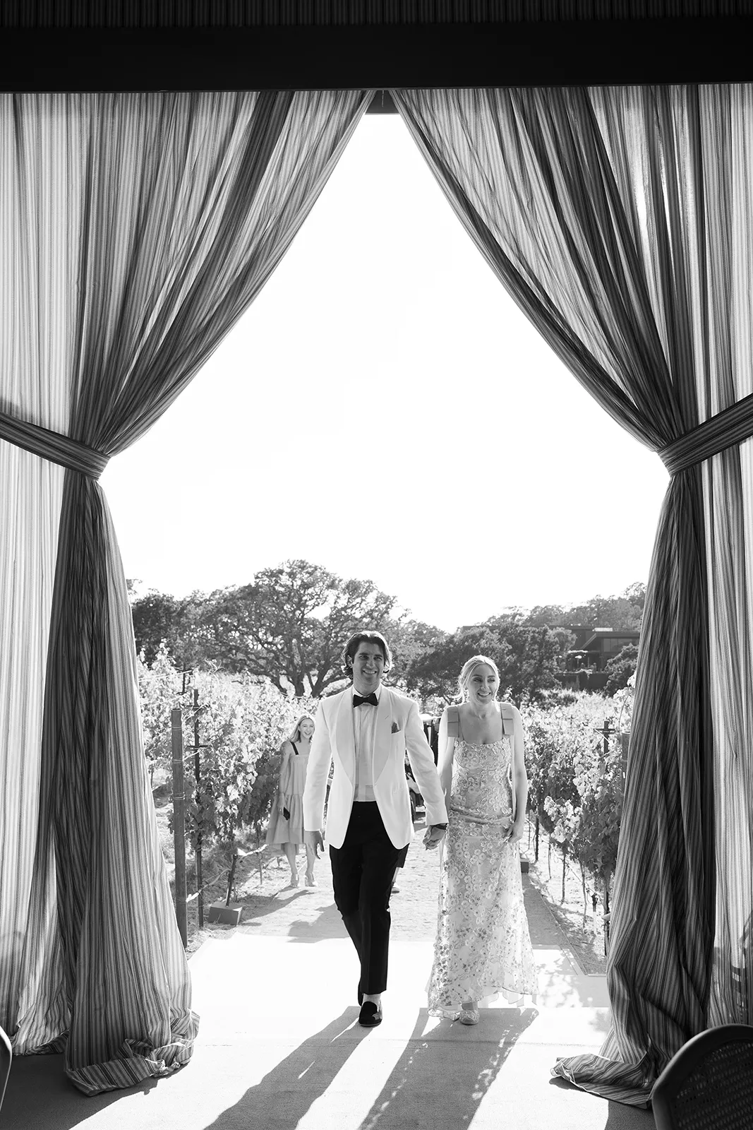 Black-and-white image of a couple walking hand in hand through sheer draped curtains toward a vineyard ceremony at Montage Healdsburg