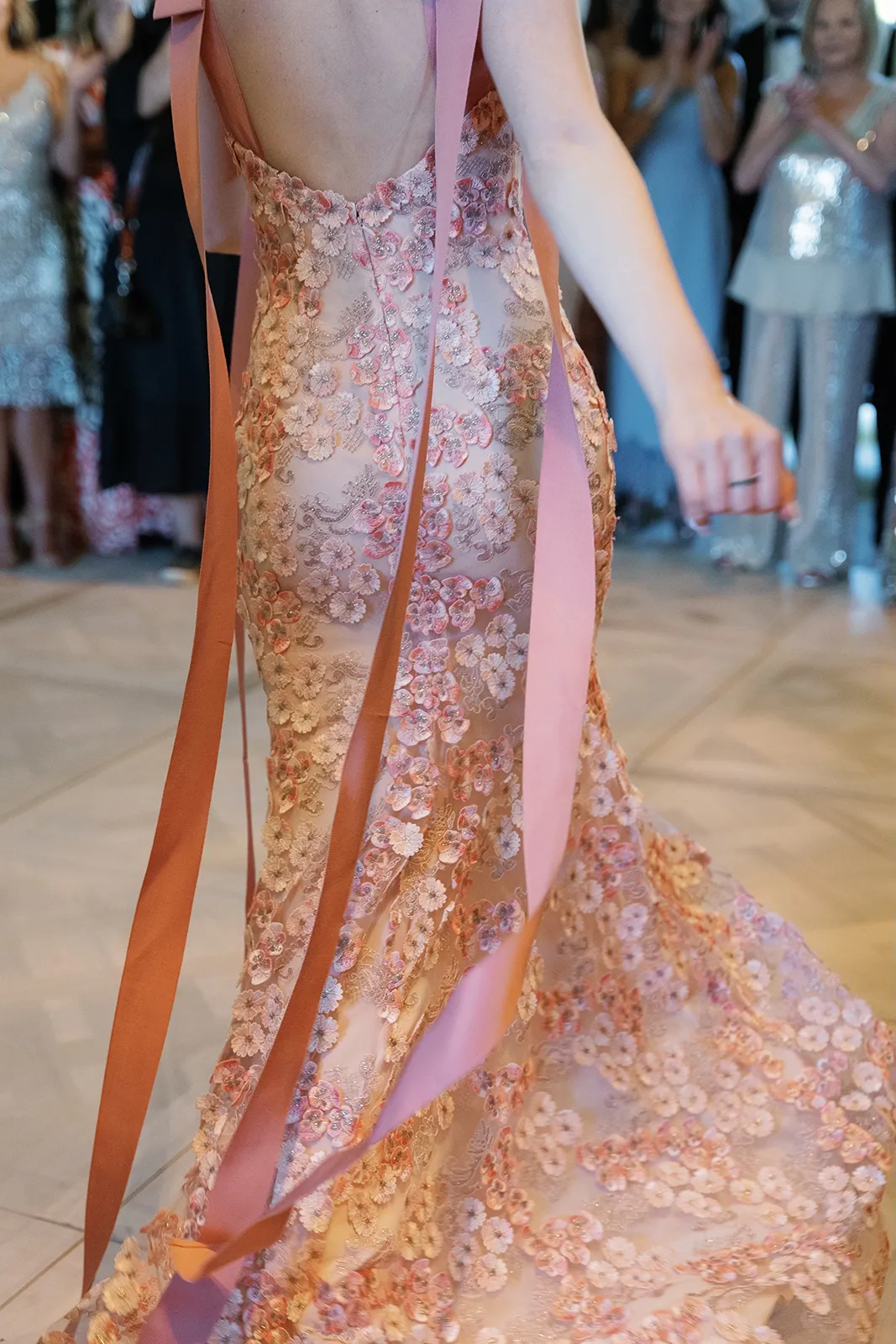 Close-up of a bride dancing at her reception in a blush floral gown with flowing ribbon details, surrounded by applauding guests