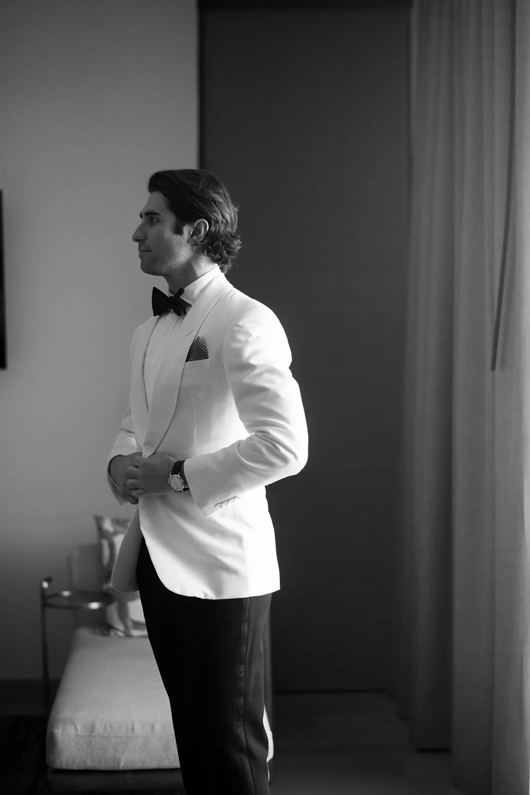 Black-and-white portrait of a groom adjusting his white tuxedo jacket inside a suite