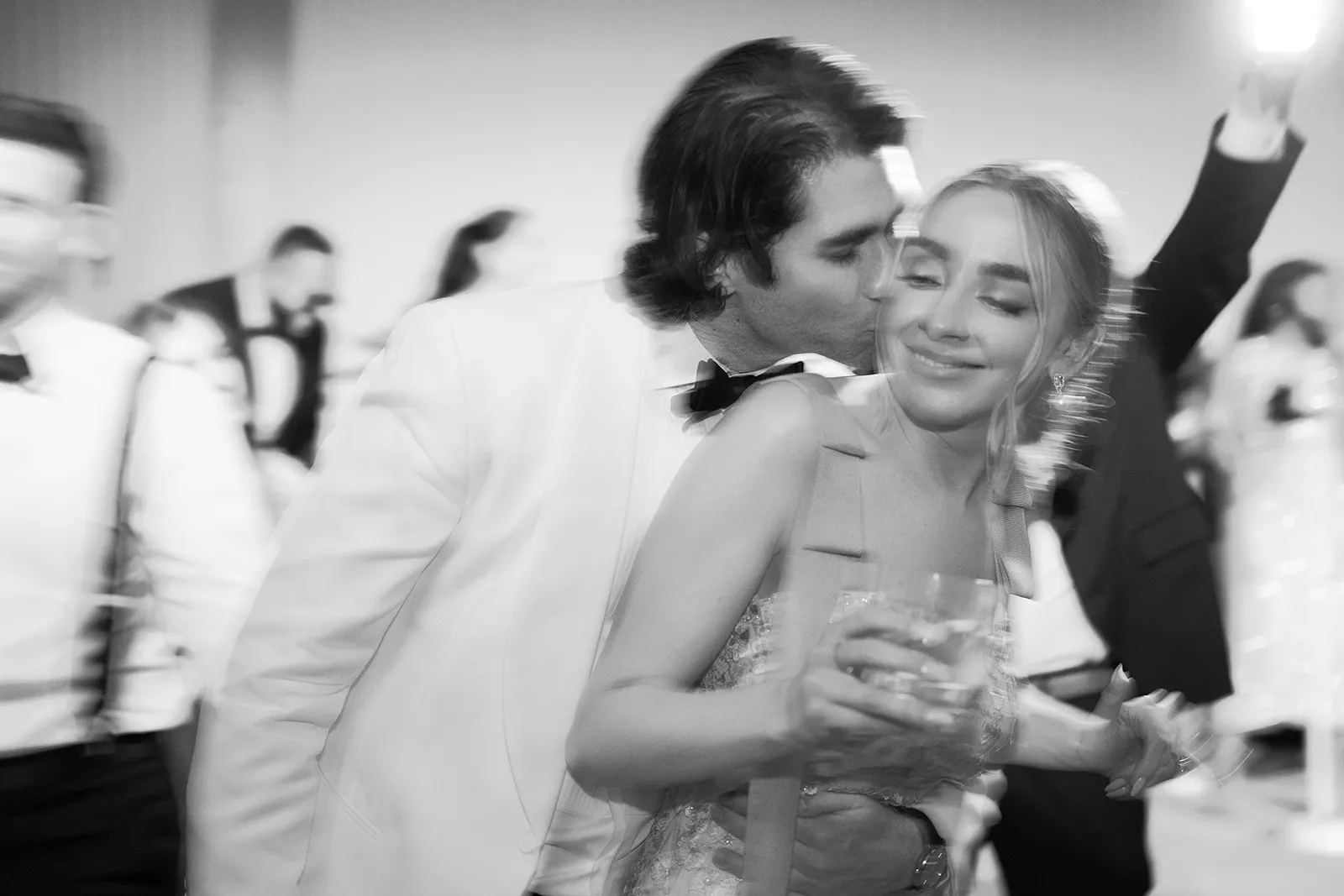Black-and-white image of the groom kissing the bride on the cheek as they dance together at their wedding reception