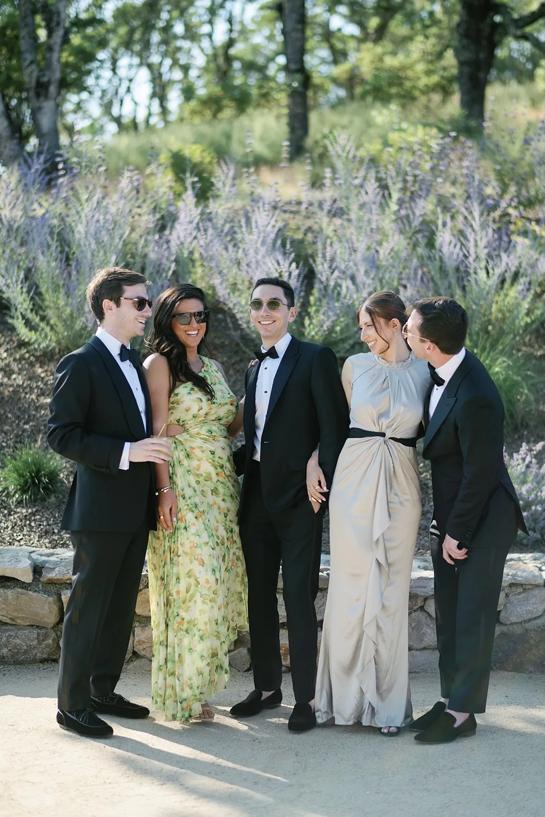 Wedding party standing together outdoors, laughing and posing in formal attire among greenery