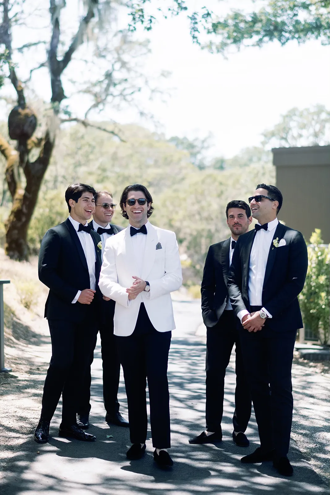 Groom in a white dinner jacket laughing with groomsmen in black tuxedos during pre-ceremony portraits