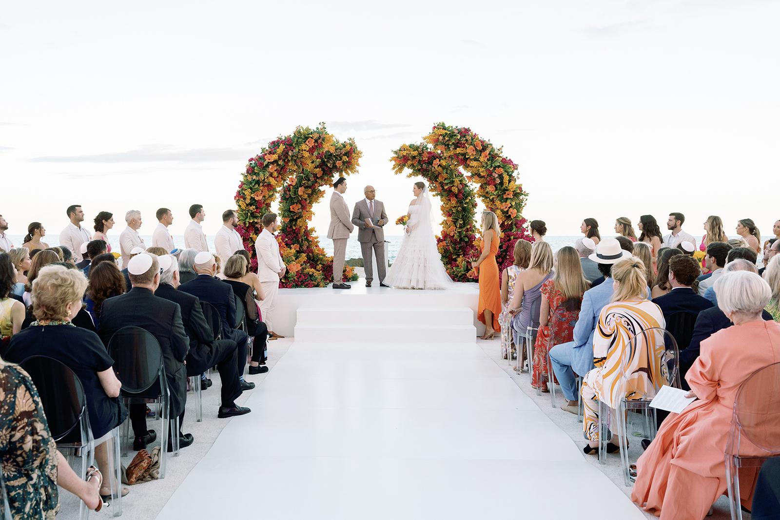 Wide view of an outdoor wedding ceremony with guests seated on both sides of a white aisle