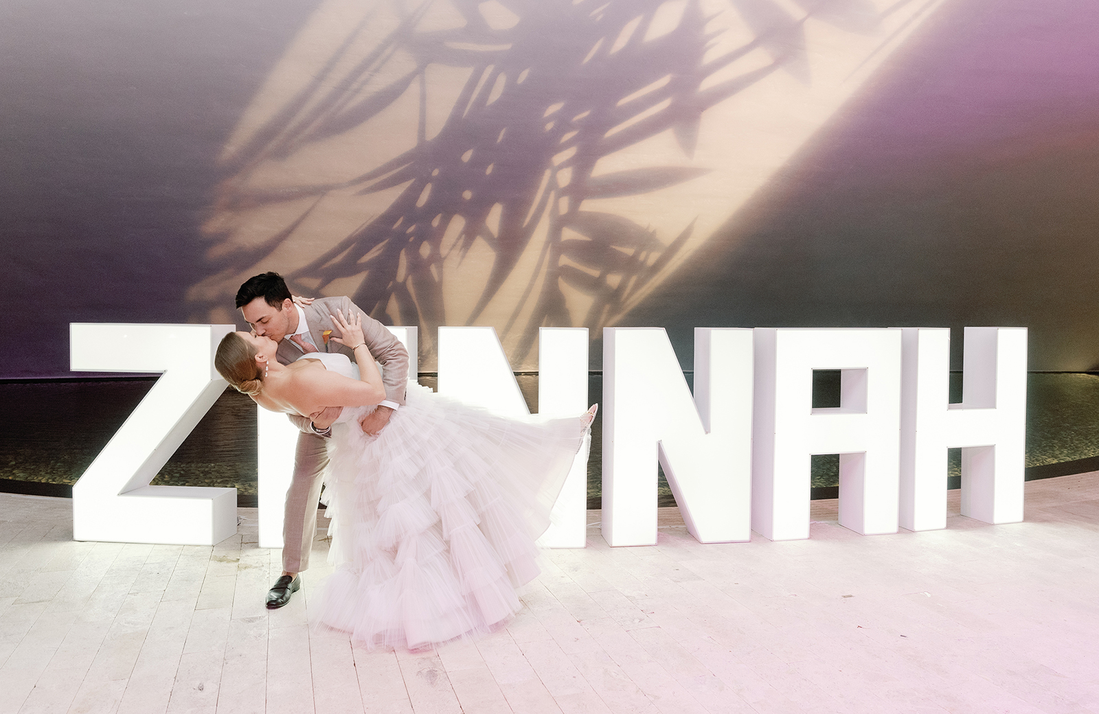 Bride and groom sharing a romantic kiss in front of a glowing modern light installation of their names