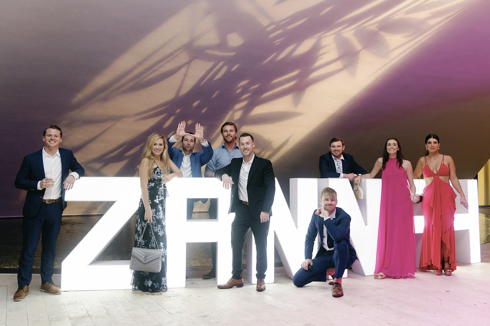 Wedding guests posing together for a group photo in front of a glowing light display of the couples' names