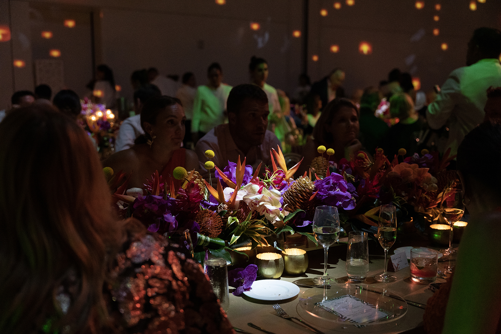 Guests seated at candlelit wedding reception table with lush tropical floral centerpiece and warm ambient lighting