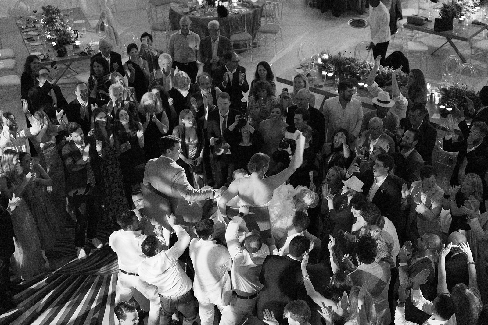 Overhead black and white photo of bride and groom lifted in chairs during a lively wedding celebration