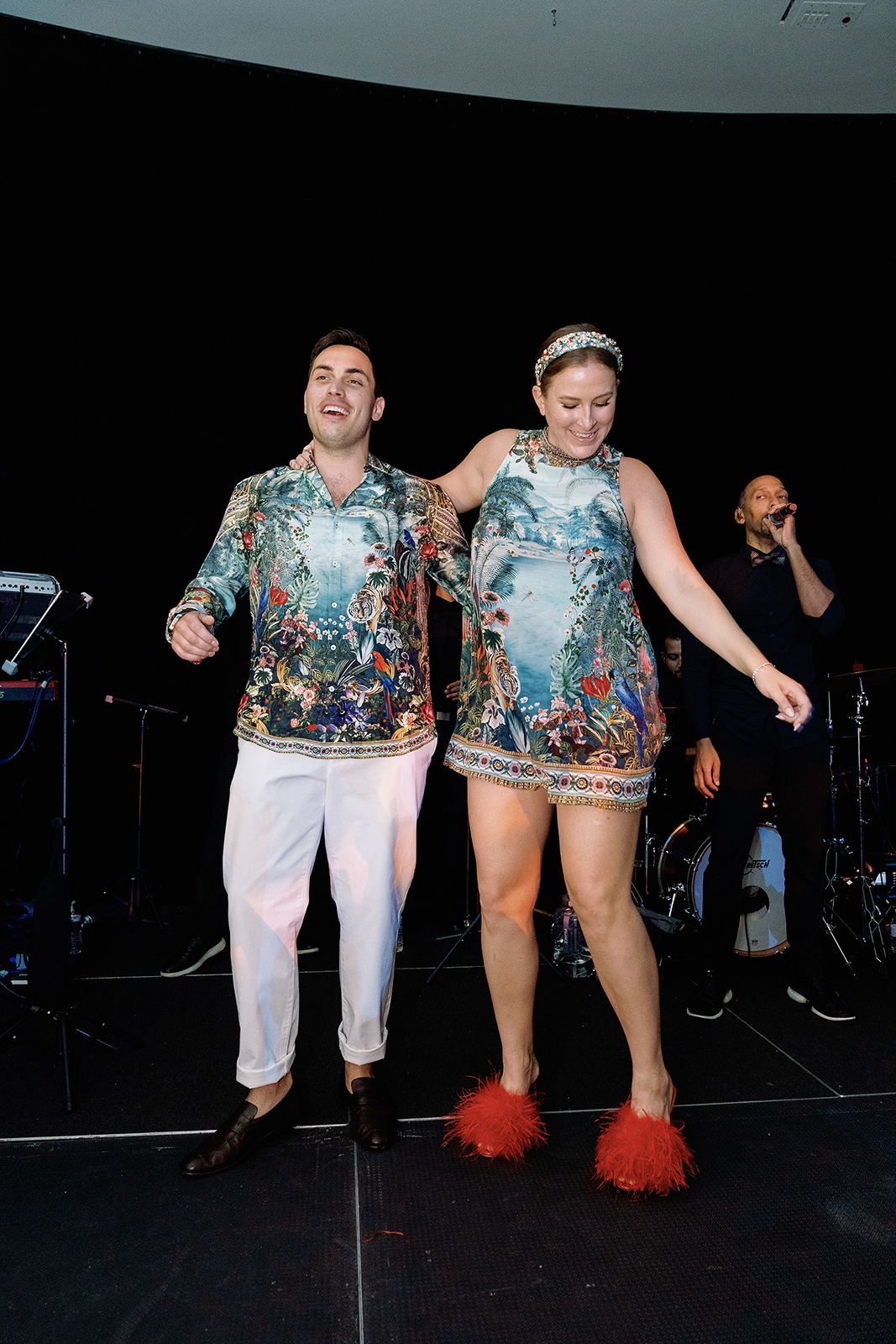 Couple dancing together on a stage during their wedding reception, wearing coordinated patterned outfits and smiling under live band lighting