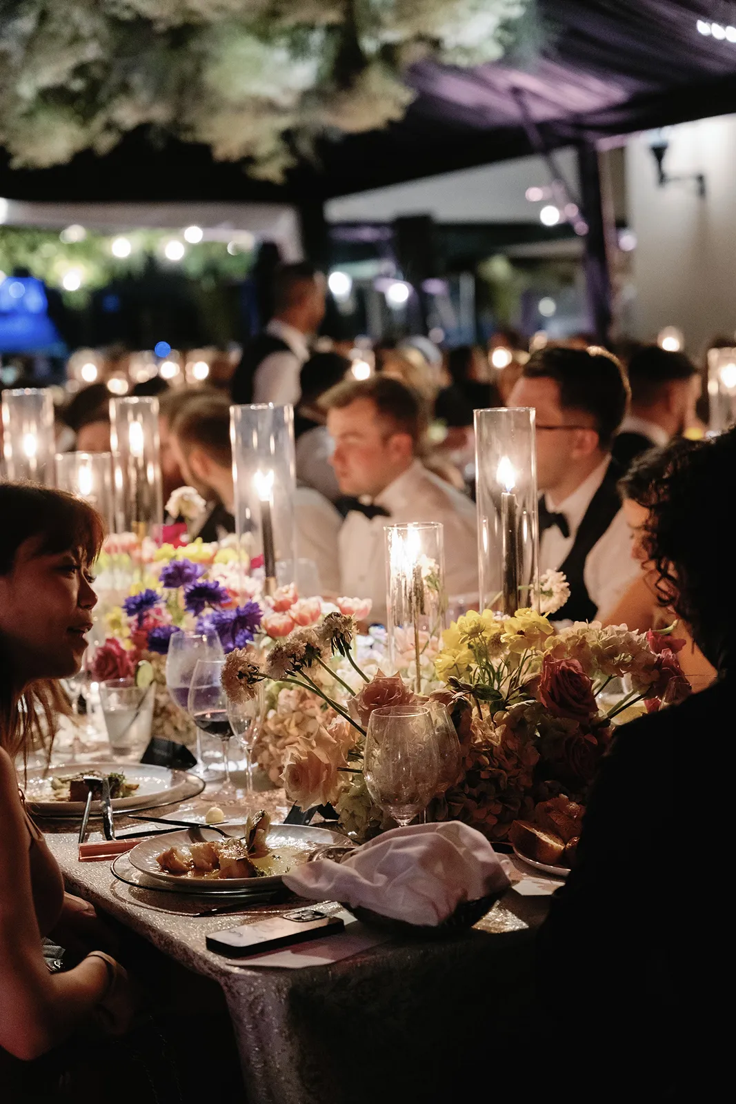 Candlelit wedding reception table with guests dining, floral centerpieces, and tall glass candles