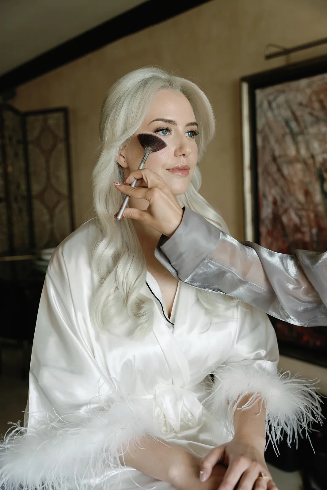 Bride getting ready as makeup artist applies blush while she wears a white robe