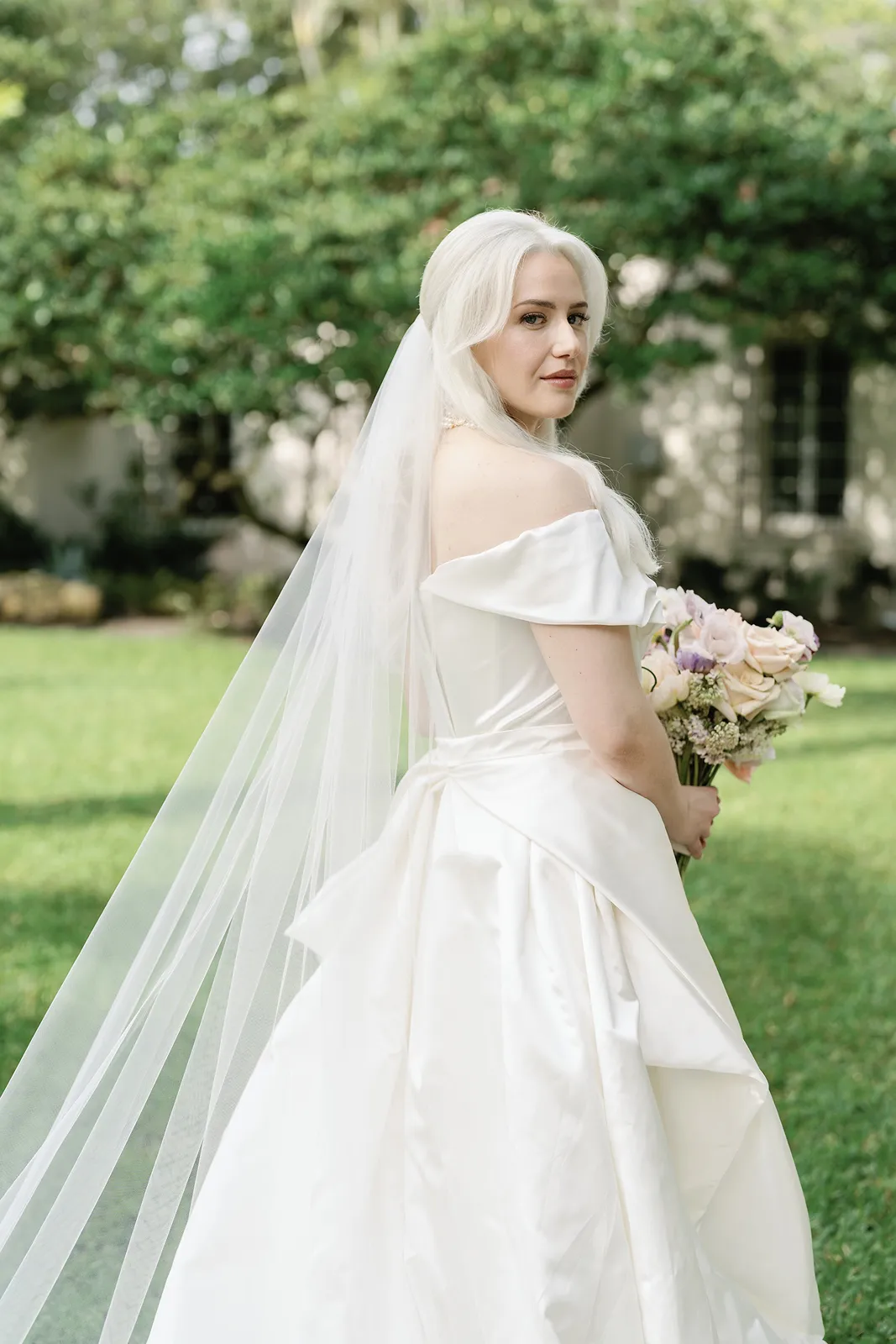 Bride in an off-the-shoulder wedding dress holding a bouquet and looking back over her shoulder outdoors