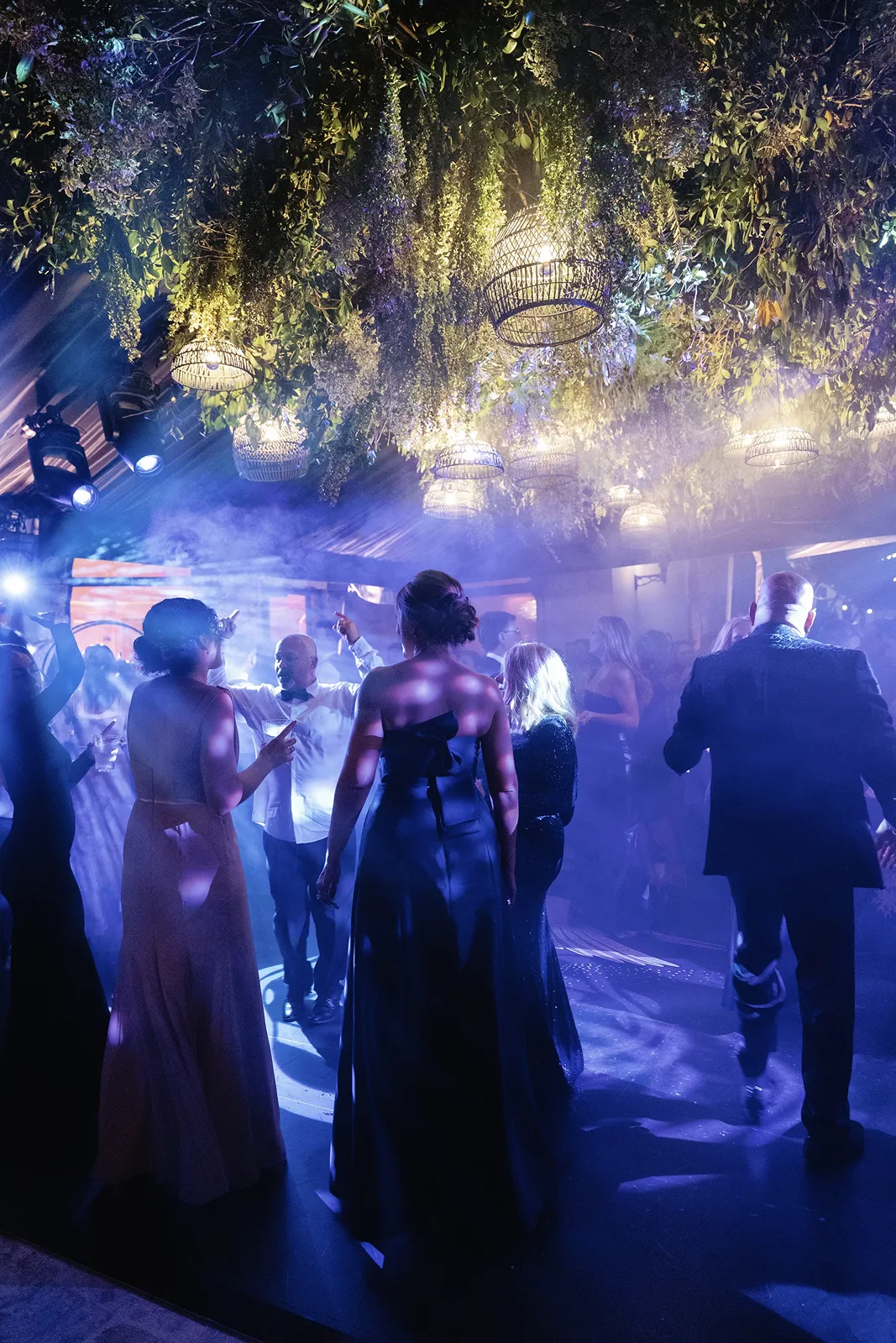 Wedding guests dancing beneath hanging greenery with colorful lighting and a festive atmosphere
