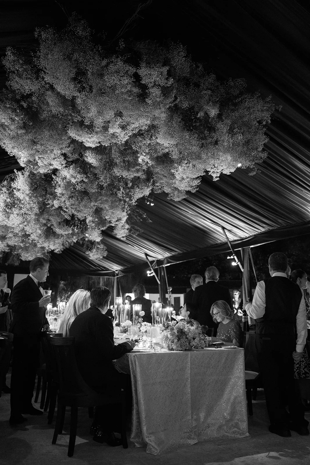 Black and white wedding reception under floral ceiling with guests dining at candlelit tables