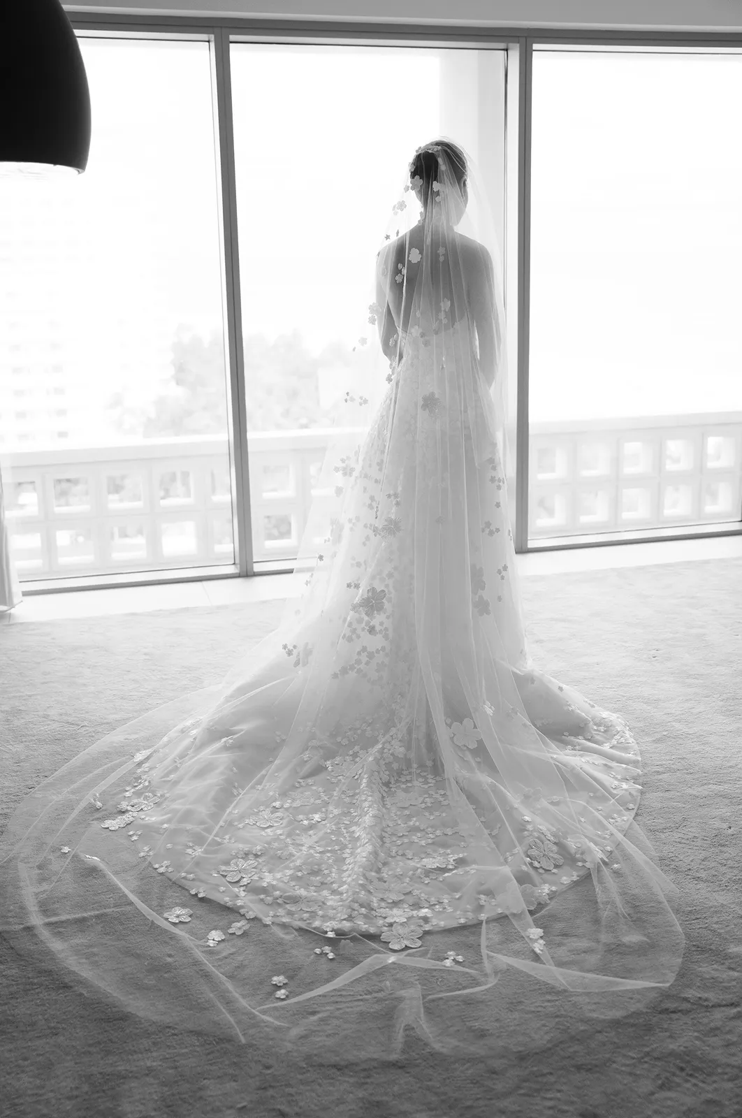 Bride standing in front of large windows wearing a lace wedding gown and cathedral-length veil