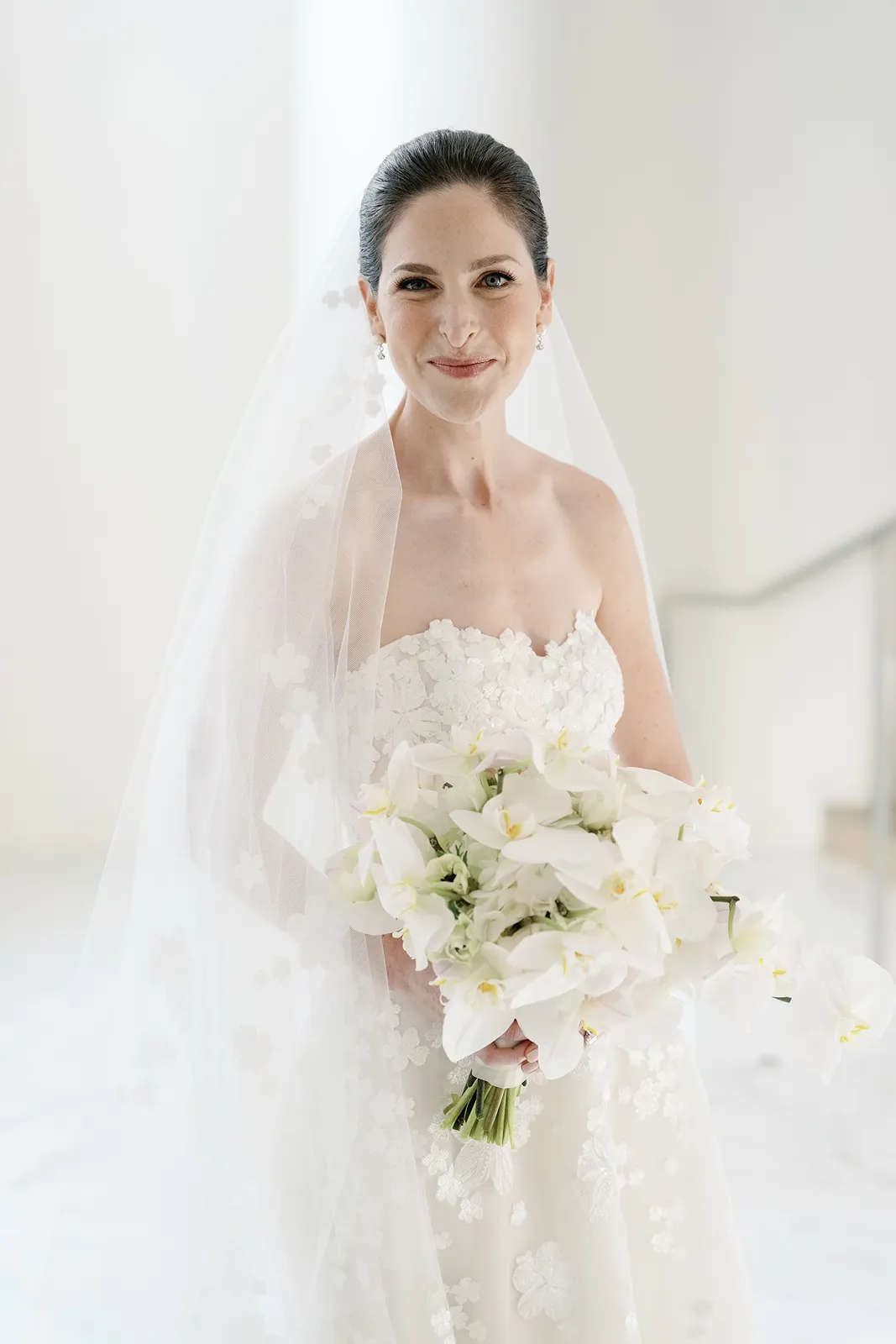 Bride smiling softly while holding a white orchid bouquet during a bright bridal portrait