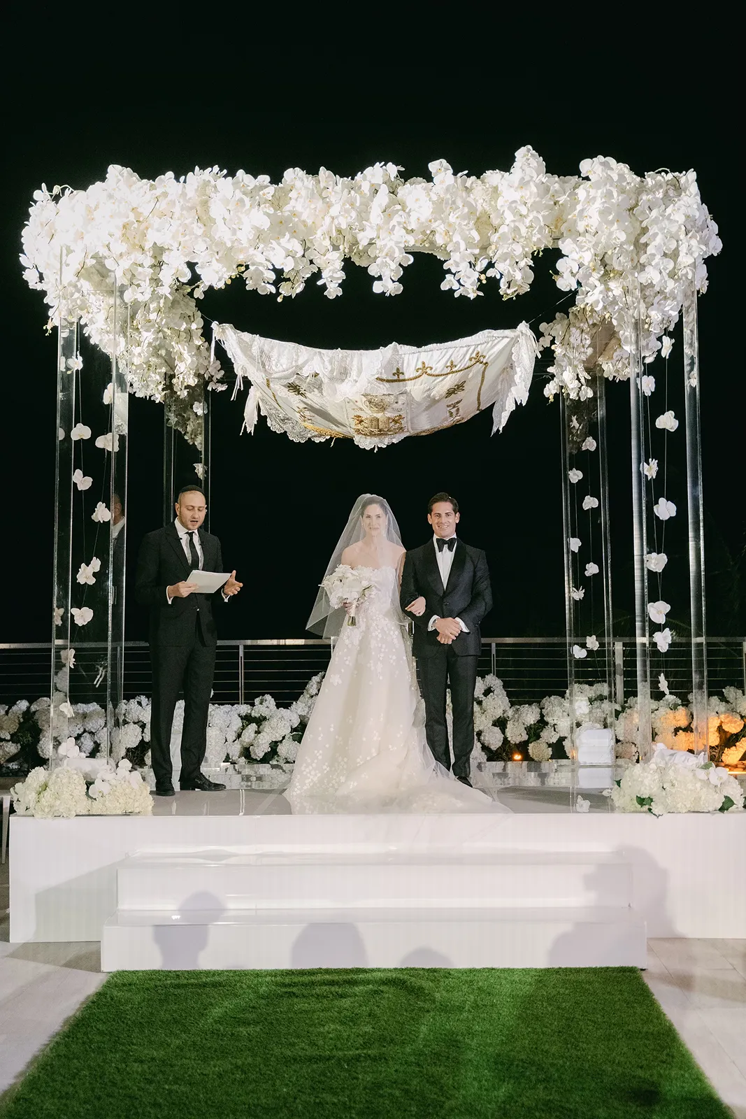 Bride and groom standing beneath a lush floral chuppah during an elegant evening wedding ceremony