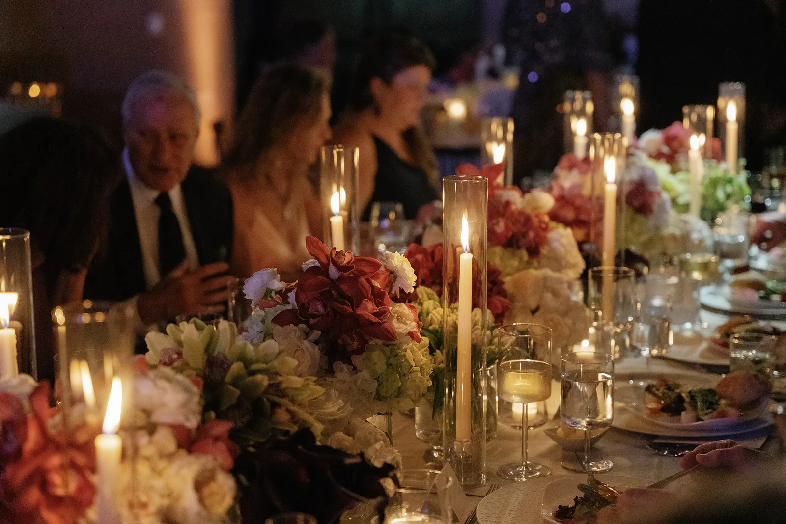 Candlelit wedding dinner table with lush floral arrangements and guests seated together in warm evening light