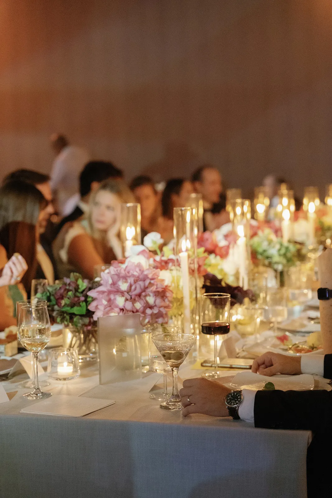 Guests seated at a candlelit wedding dinner table surrounded by soft florals and warm ambient light