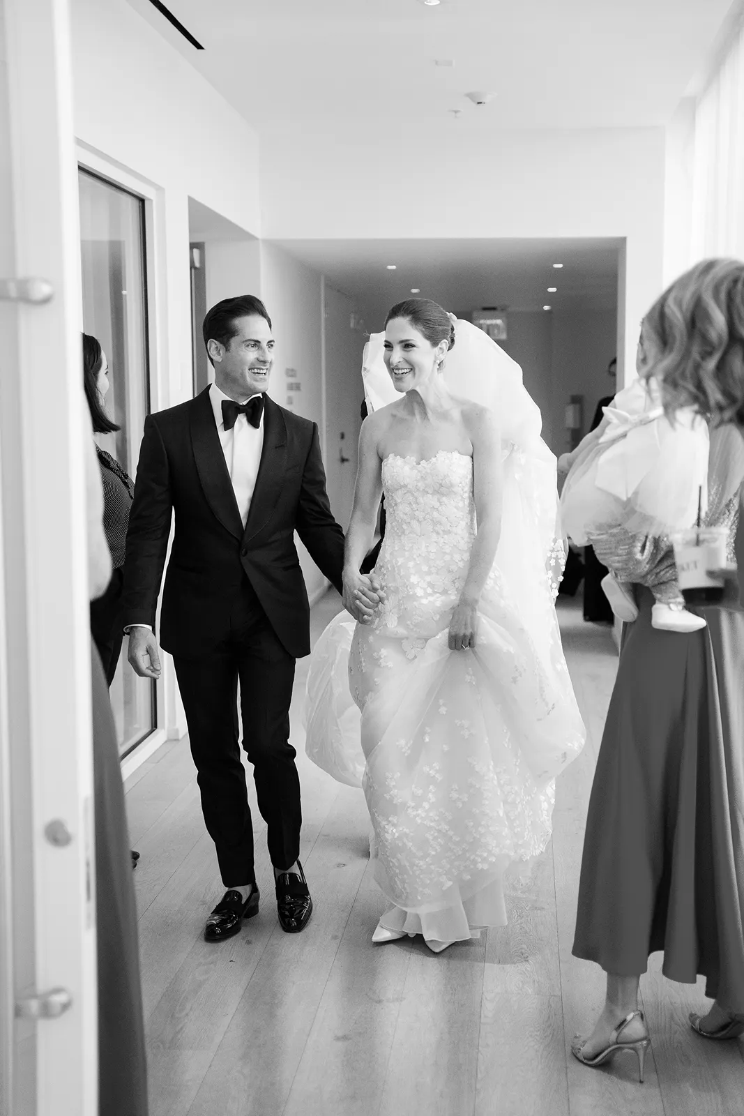 Bride and groom walking hand in hand through a bright hallway surrounded by family and friends