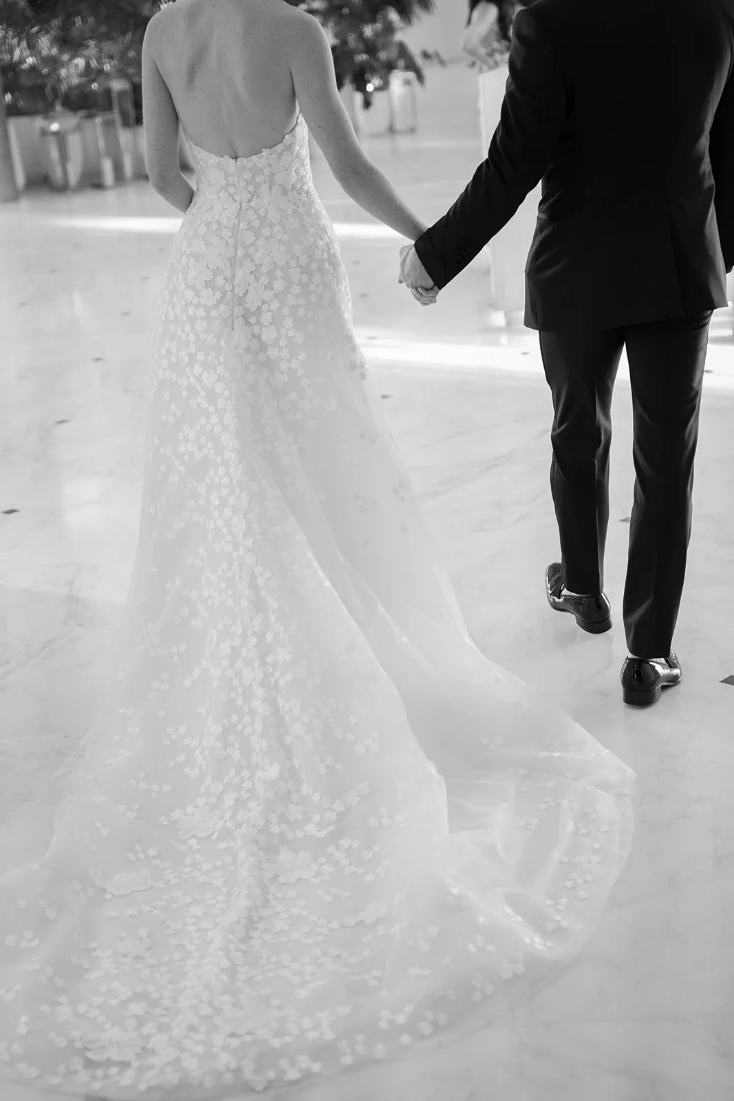 Bride and groom walking hand in hand from behind, the bride’s lace gown trailing softly along the floor