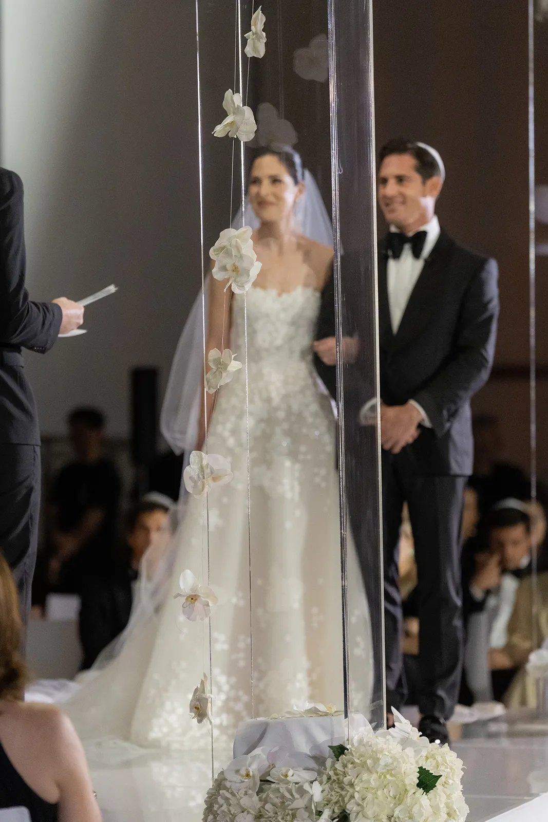 Bride and groom standing together during the ceremony, seen through clear acrylic columns with hanging floral details