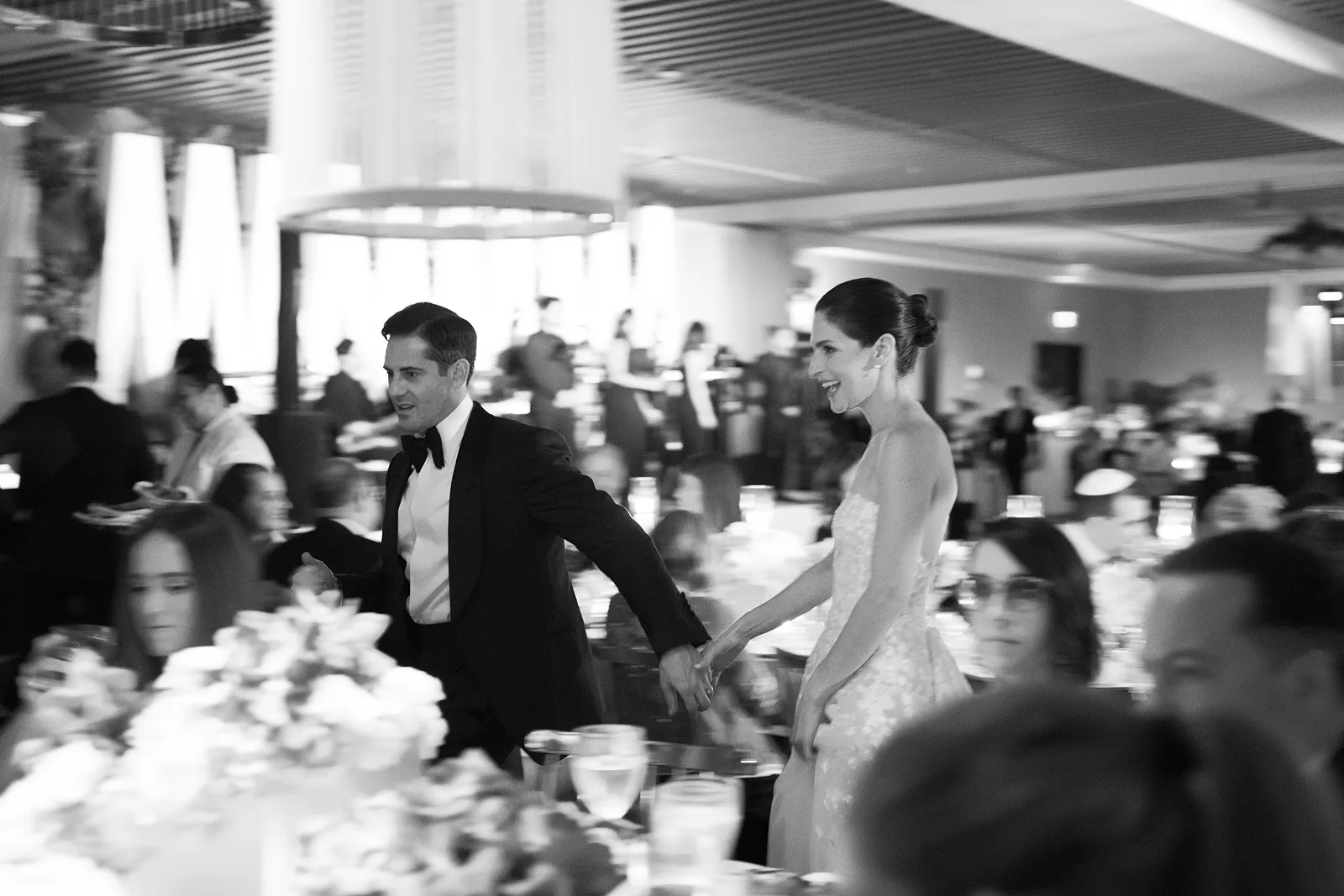 Bride and groom holding hands as they walk through a lively reception space filled with guests