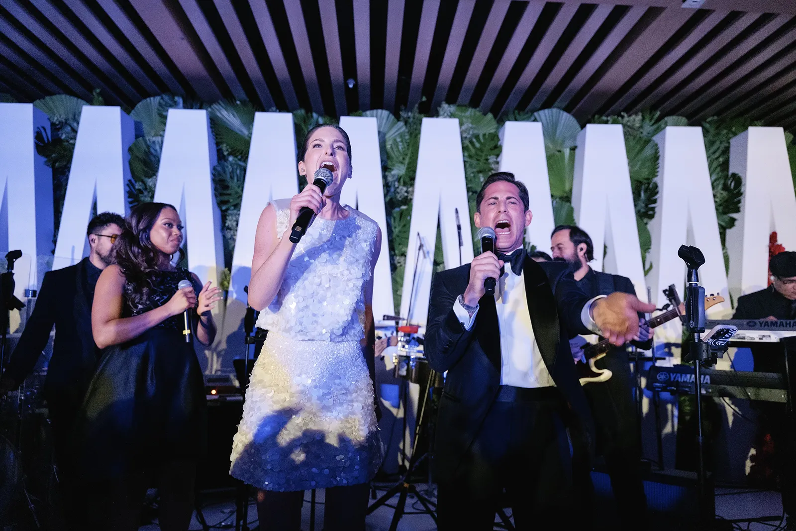 Bride and groom singing into microphones on stage with the live band during the reception