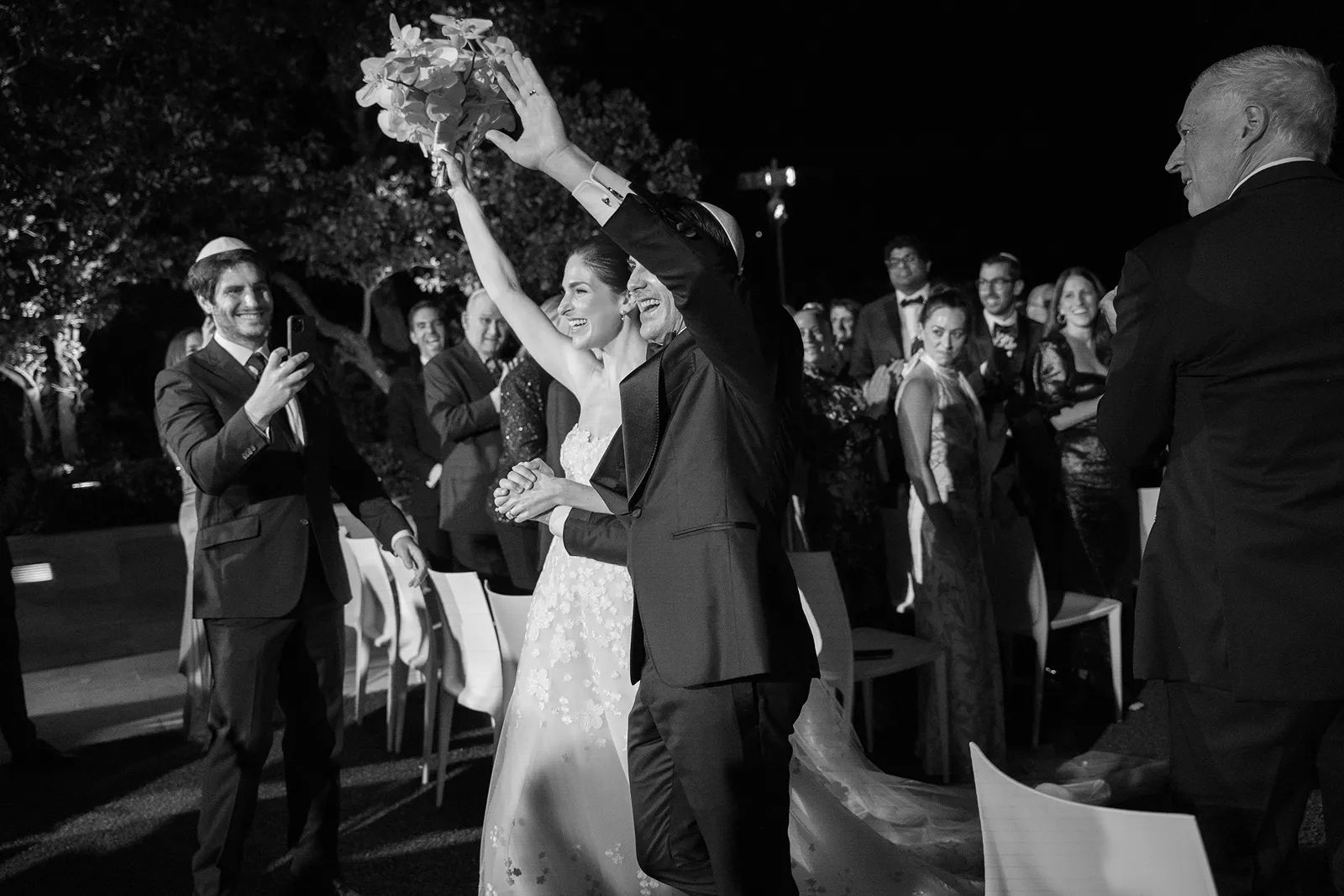 Newlyweds celebrating as they walk back up the aisle, raising the bouquet while guests cheer around them
