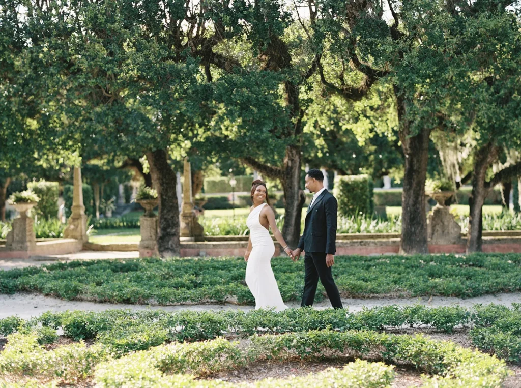 couple walking through vizcaya gardens as bride looks back at her husband to be