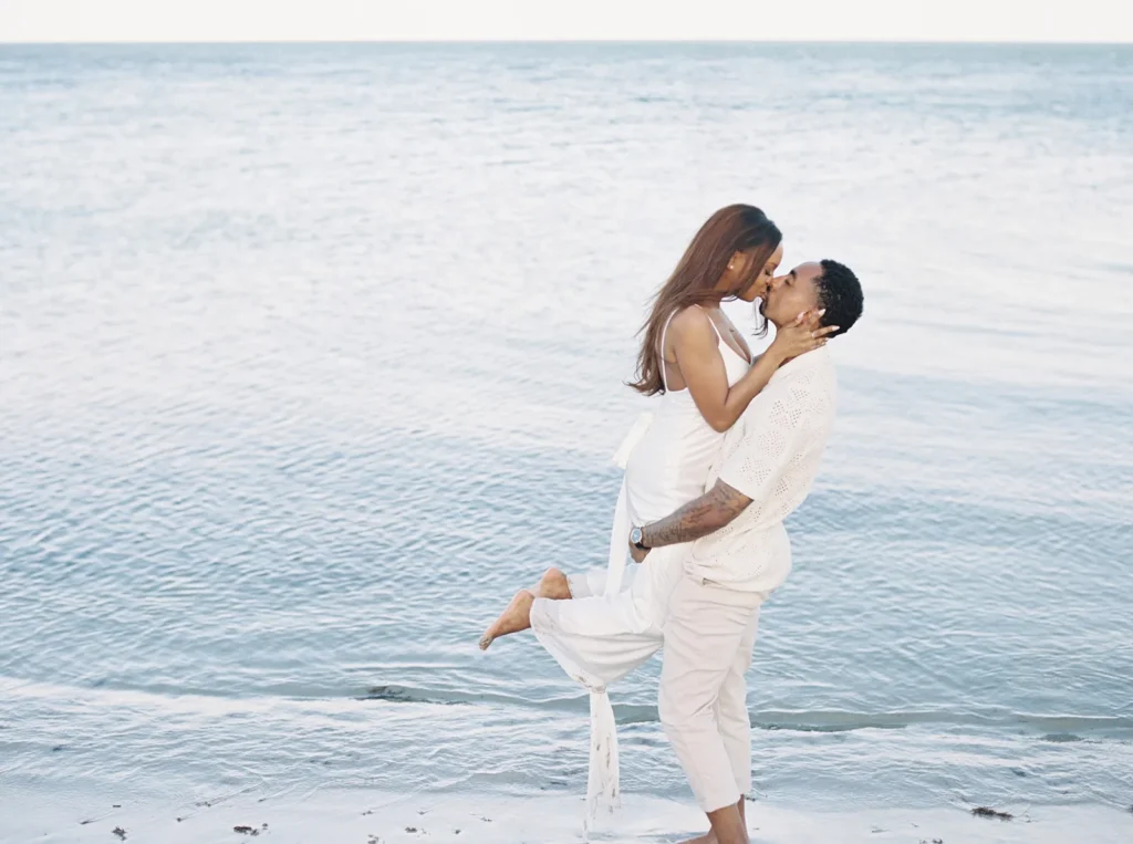 groom carrying his fiancé as they kiss with the ocean behind them