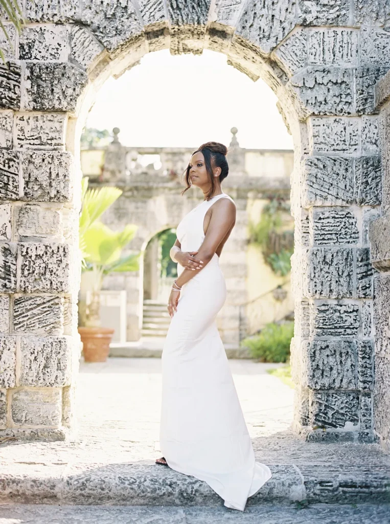 bride in white silk dress under an archway with sun shining through