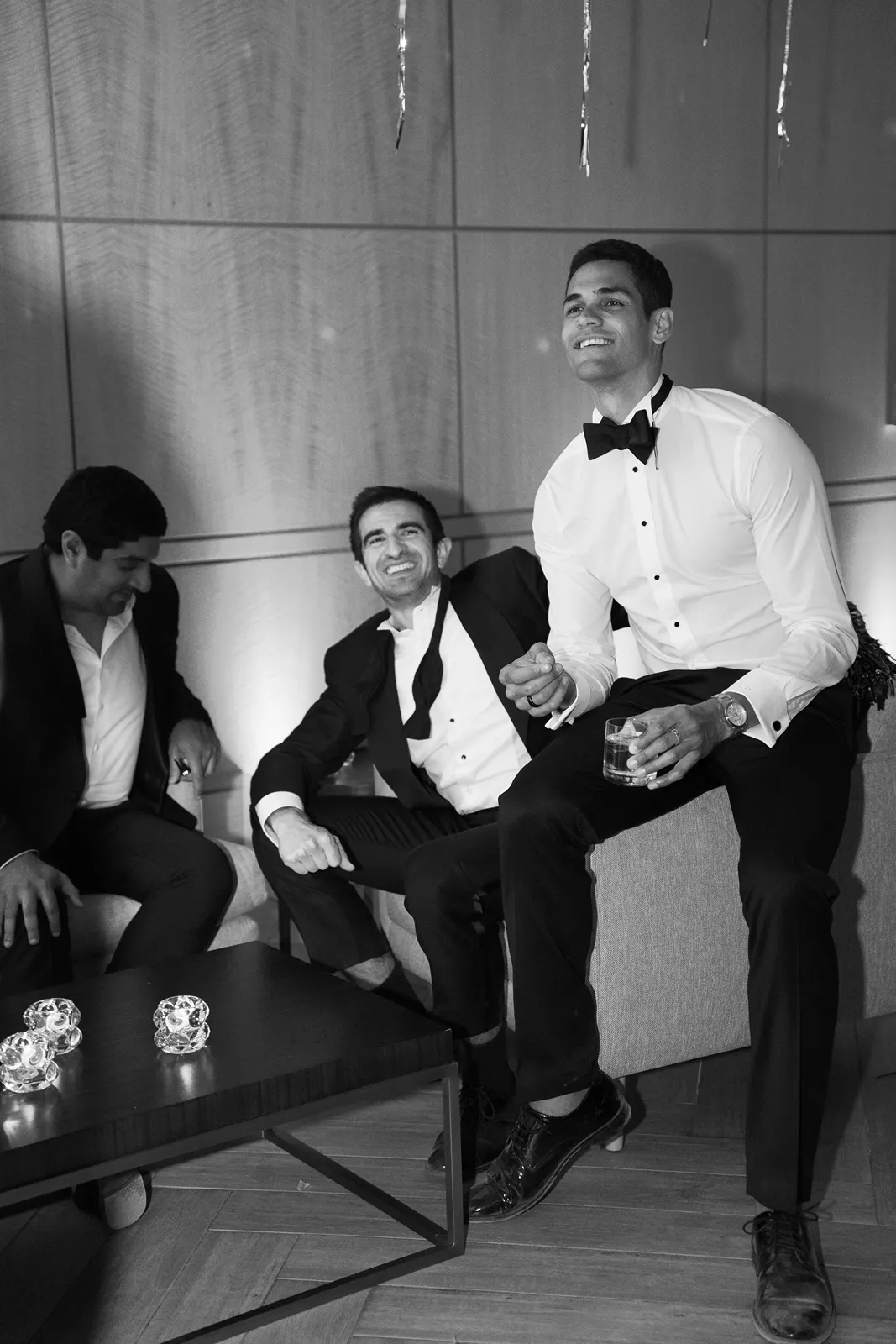 Friends relax on lounge seating, dressed in black tie attire, enjoying cocktails during a wedding after-party