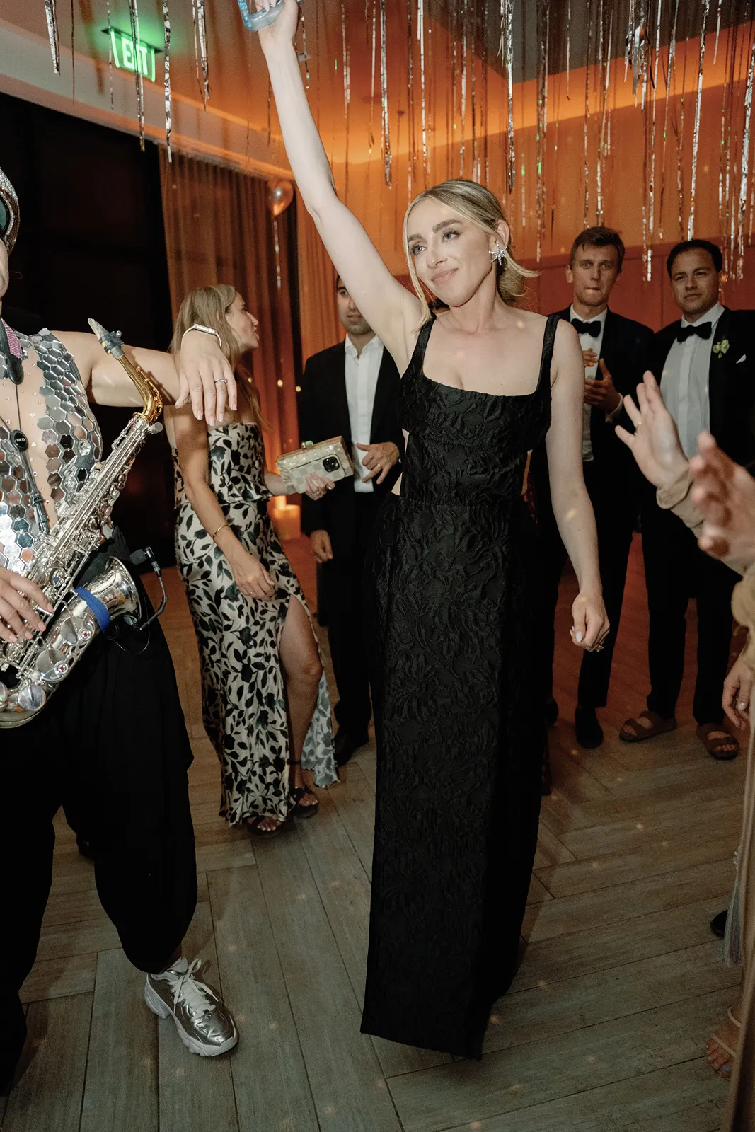 Bride raises a hand while dancing beside a live saxophonist during a high-energy after party