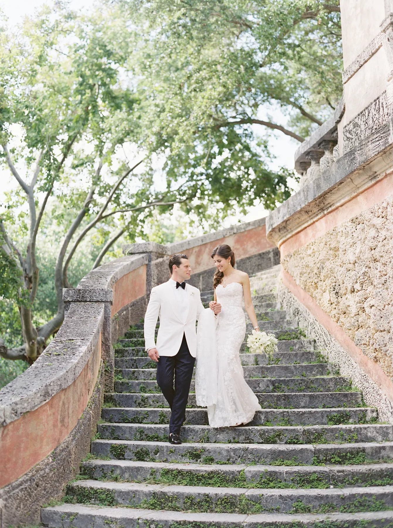 Bride and groom walking hand in hand down curved stone stairs surrounded by greenery