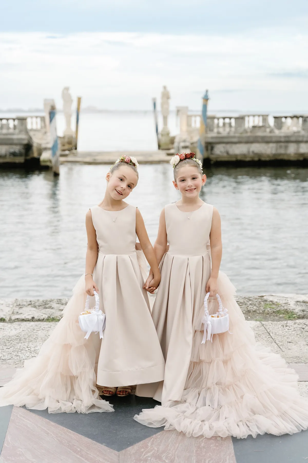 Two flower girls holding baskets and smiling by the water before the ceremony